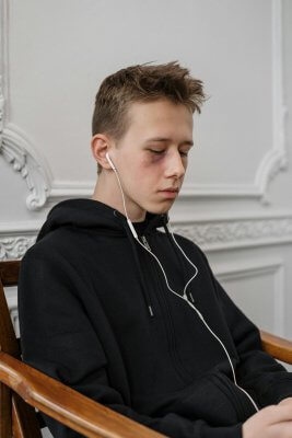 A teenager wearing a black hoodie listens to music with earphones while sitting indoors.